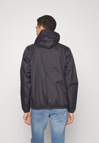 Jachetă neagră cu glugă, cu tiv elastic, din material neted de nylon, cu detalii minime. Purtați-o cu blugi bleu deschis, arătând o vedere din spate casual.