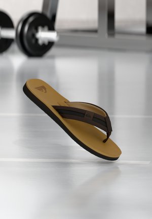 Brun og svart flip-flops sandal som svever over et reflekterende gulv med uklare treningsvekter i bakgrunnen.