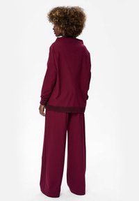 Vrouw met krullend haar die een losse, lange mouwen bordeauxrode top en bijpassende wijde broek draagt, rechtopstaand met haar rug naar de kijker gekeerd.