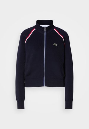 Navy zip-up jacket in soft material with red and white accent stripes. Features a high collar and Lacoste logo on the chest.