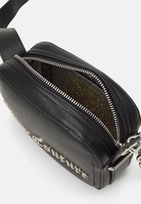 Svart syntetisk axelväska med en strukturerad yta, silverf ärgad dragkedja och metallisk text på framsidan. Öppen dragkedja visar ett mönstrat inre.