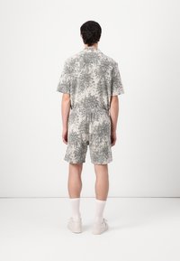 Kortermet, krage jumpsuit i lysegrå med et mørkegrått abstrakt mønster. Elastisk midje, shorts og bakside passform. Enkle hvite sneakers.