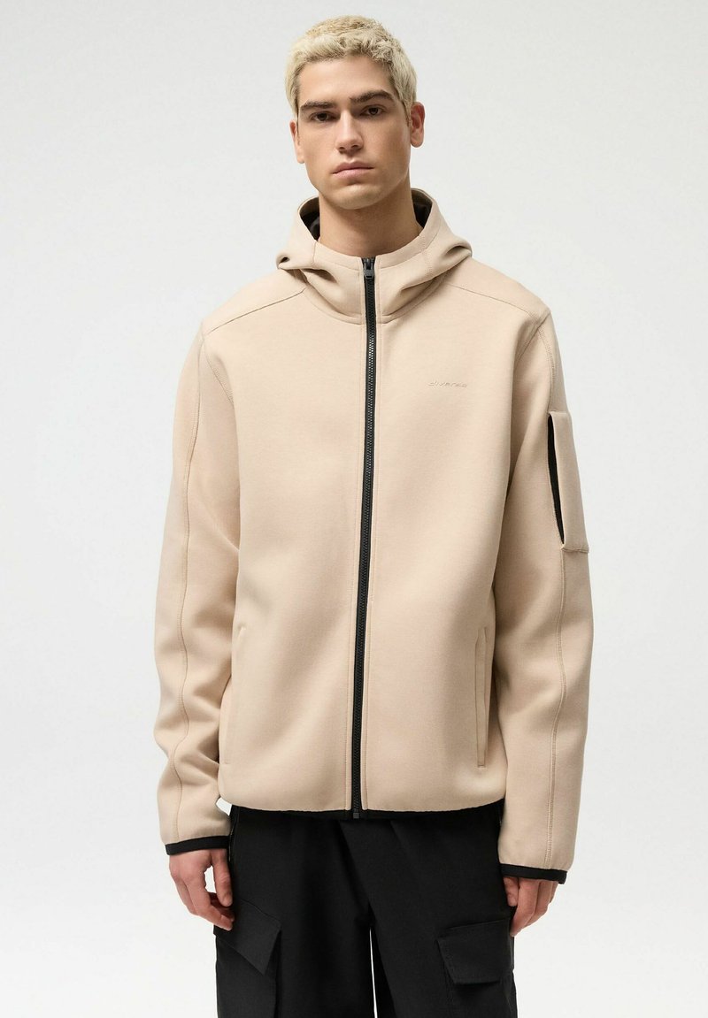 Młoda osoba z krótkimi blond włosami, ubrana w beżową bluzę z kapturem i zamkiem błyskawicznym, z kieszenią na lewym rękawie.