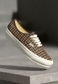 Brązowe trampki w wzór houndstooth z płótna, z białymi sznurówkami i gumową podeszwą, z wyściełanym kołnierzem oraz metką z logo po boku.