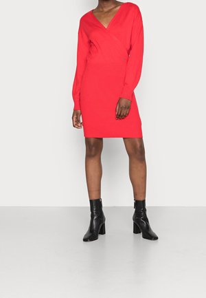 Personne portant une robe rouge à manches longues avec un col en V et des bottines noires, debout devant un fond clair uni.