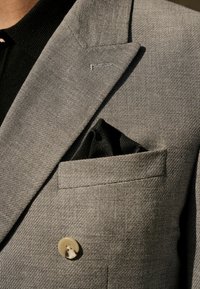 Blazer texturé gris avec un bouton clair unique et un poche noire dans la poche poitrine. Coupe ajustée avec des revers nets.