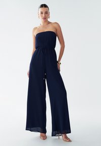 Temno modra breznaramnična jumpsuit z nabrano korzeto, sproščenim krojem in širokimi hlačnicami. Opremljeno z vrvico v pasu in mehko teksturo tkanine.