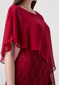 Przezroczysta czerwona szyfonowa nakładka o płynącym kroju, nałożona na dopasowaną koronkową spódnicę z motywem kwiatowym i cekinami.
