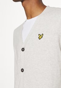 Hellgrauer Cardigan mit tiefem V-Ausschnitt, ausgestattet mit schwarzen Knöpfen und einem kleinen gelben gestickten Logo eines Vogels auf der linken Brust.
