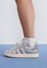 Gray suede sneaker with light gray stripes, white laces, and a rubber sole. Worn with a white ankle sock, set against a blue background.