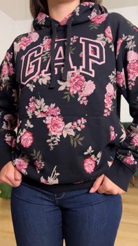 Μπλε φούτερ με κουκούλα με ροζ ανθισμένο σχέδιο και μεγάλο λογότυπο GAP, φορεμένο με σκούρο μπλε τζιν, τα χέρια στις τσέπες.