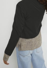 Mustat lyhyt takkimekko pitkähihaisilla, jossa on teksturoitu beige neulekangas vyötäröllä ja napit hihansuissa, yhdistettynä vaaleansineisiin farkkuihin.