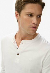 Weißes langärmliges Henley-Shirt aus Baumwolle, mit einem Dreiknopfverschluss aus braunen Knöpfen und einem dezenten Logo auf der Brust.