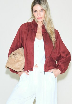 Woman with long blonde hair wearing a burgundy zip-up jacket over a white buttoned top, white pants, holding a tan suede clutch purse.