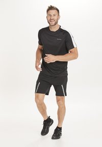 Musta urheilupaita valkoisilla hartiakohdistuksilla, yhdistettynä mustiin shortseihin, joissa on heijastavat raidat sivuilla. Mustat lenkkarit viimeistelevät asun.