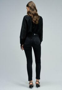 Blusa negra transparente, jeans ajustados negros de cintura alta y tacones negros con tiras. Textura suave con sutiles detalles en los bolsillos de los jeans.