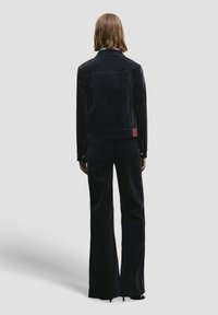 Giacca in velluto nero con taglio aderente, cuciture visibili e una piccola toppa con logo sul retro. Abbinata a pantaloni svasati coordinati.