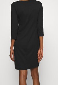 Robe noire à manches trois-quarts, forme ajustée, encolure ras du cou et tissu texturé. L'arrière présente une couture centrale et un ourlet juste au-dessus du genou.