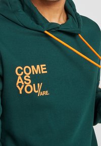 Grønblå hættetrøje med orange snøre. Har teksten "COME AS YOU/ARE." trykt med fed skrift på forsiden. Blødt stof, afslappet pasform.