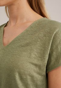 Olivově zelený krátký rukáv top s výstřihem do V, vyrobený z měkkého, texturovaného materiálu a uvolněného střihu. Výstřih je navržen pro pohodlí.