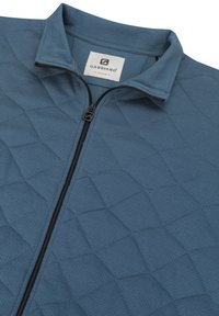 Chaqueta de forro polar azul con textura acolchada, un cuello alto y una cremallera en la parte delantera con herrajes negros. Etiqueta de la marca visible en el interior del cuello.