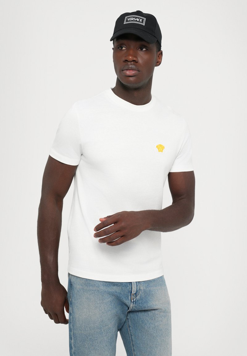 Vit t-shirt med kort ärm som har en liten gul logo på bröstet, ihopparad med ljusblå denimjeans. Tyget verkar slätt.