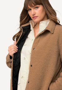 Brun fleecemantel med en texturerad yta, med en svart quiltad foder och tryckknappar. Under har den en vit skjorta med blomsterbroderi.