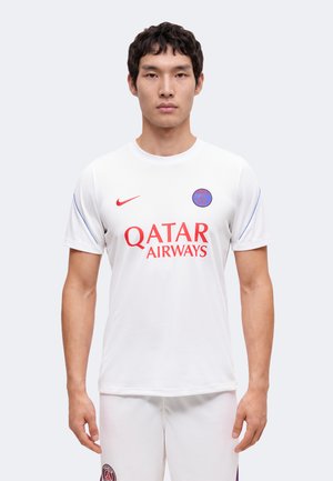 Jeune homme debout, faisant face, portant un maillot de football blanc du Paris Saint-Germain avec un logo Qatar Airways rouge et un symbole Nike.