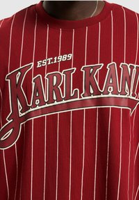 T-shirt rossa in cotone con strisce bianche verticali, caratterizzata da una grande stampa grafica di "KARL KANI" e "EST.1989" in colori a contrasto.