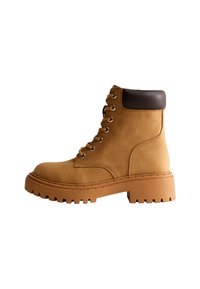 Stivale alla caviglia in suede tan con colletto in pelle marrone, lacci sul davanti, punta rotonda e suola in gomma spessa con un motivo a trama testurizzata.