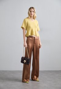 Suéter corto amarillo y esponjoso, pantalones de satén marrón de pierna ancha, zapatos verdes de punta, y un bolso negro con estampado y cadena dorada.