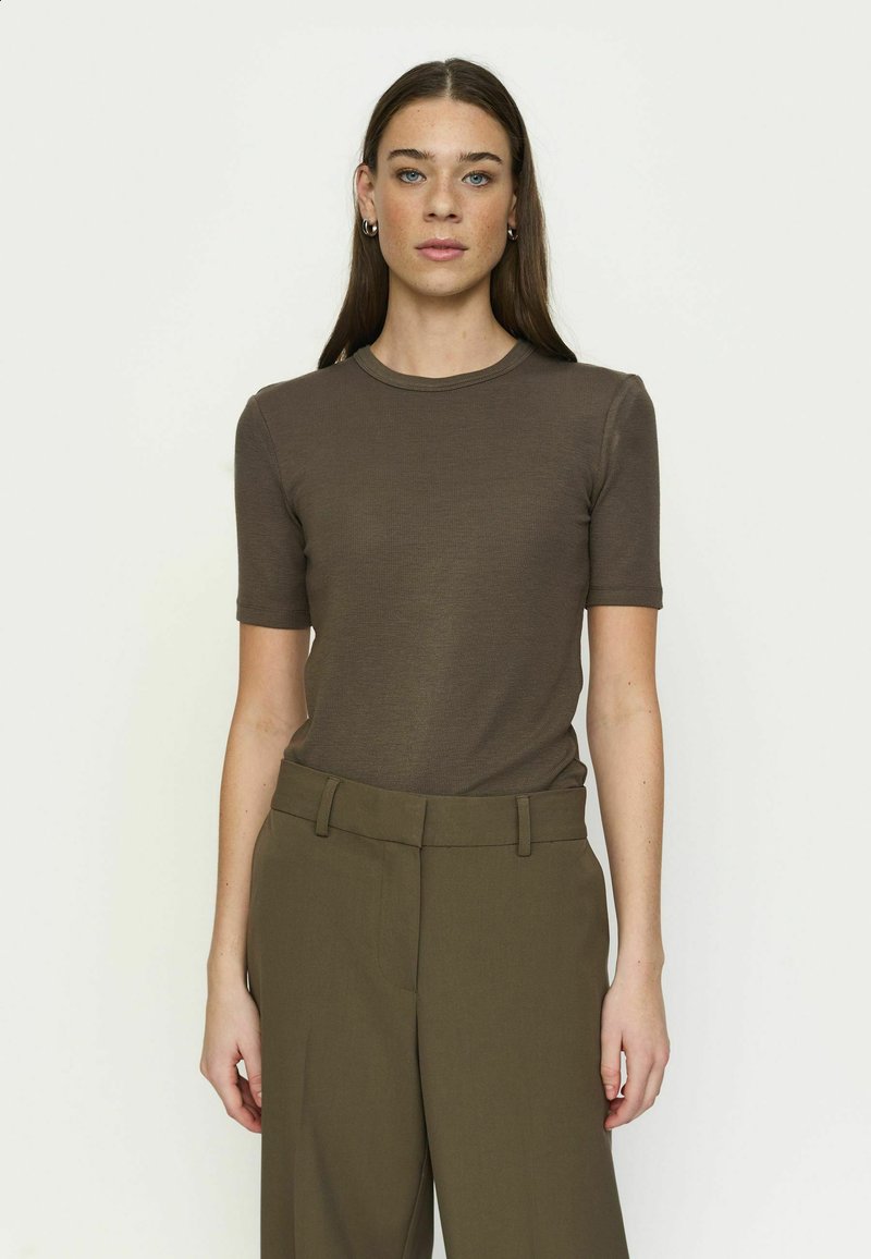 Femme aux cheveux bruns et lisses porte un t-shirt marron ajusté à manches courtes et un pantalon olive taille haute, debout devant un fond uni.