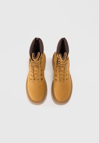 Gule støvler med en glat overflade, brun læderkrave og runde snørebånd. Gummisål og forstærket tå for holdbarhed.