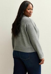 Maglione grigio a maglia con una vestibilità rilassata, orlo e polsini a coste, indossato su jeans blu scuro. Capelli lunghi e mossi che scendono sulla spalla.