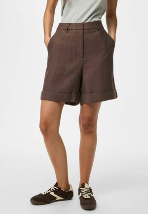 Nő barna felhajtott szárú rövidnadrágot, szürke ujjatlan felsőt és barna sportcipőt visel fehér fűzőkkel, zsebre tett kézzel áll.