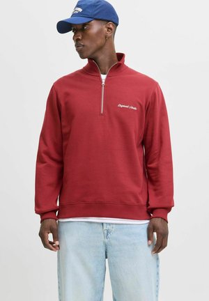 Sudadera roja con media cremallera y puños y dobladillo acanalados. Presenta un logo bordado en el pecho. Lleva puesta sobre una camisa blanca con capas y jeans de color azul claro.