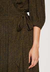 Main droite reposant à côté d'une taille nouée avec un nœud sur une robe vert olive foncé avec un petit motif léopard noir.