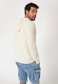 Crème-kleurige gebreide hoodie met een gestructureerd patroon, voorzien van een capuchon en een geribbelde zoom, gecombineerd met lichtblauwe cargoshorts.