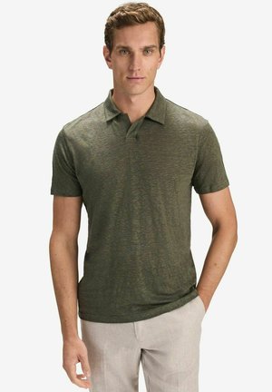 Man draagt een olijfgroen poloshirt met korte mouwen en lichtbeige broek, staat met één hand in de zak tegen een effen achtergrond.