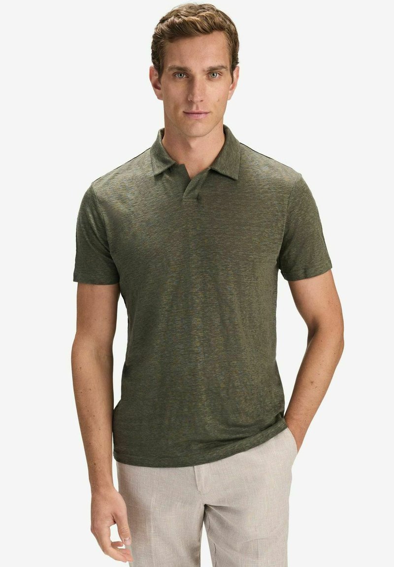 Homme portant un polo à manches courtes vert olive et un pantalon beige clair, debout avec une main dans la poche devant un fond uni.