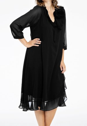 Vrouw die een zwarte, knie-lange chiffonjurk draagt met driekwartmouwen en een gerimpelde halslijn, staand met één hand op de heup.