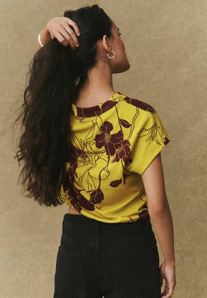 Sárga, virágmintás rövid ujjú felső bordó mintával. Fekete nadrággal párosítva, puha anyagból és laza szabásban.
