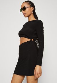 Robe noire à manches longues avec un design découpé à la taille, accentuée par des anneaux en métal. Fabriquée dans un tissu lisse et extensible.