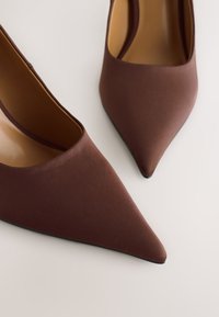 Brune højhælede sko med spids tå lavet af glat stof med en glat finish. Indersiden er tanfarvet, med en sort ydersål. Ingen synlige metaldele.