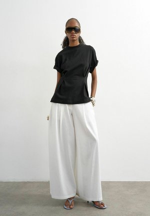 Kvinde iført sort kortærmet top, brede hvide bukser, store solbriller og sandaler, står foran en hvid væg.