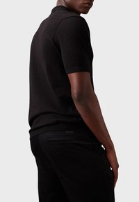 Zwarte poloshirt van gebreide stof, met korte mouwen en een klassieke kraag. De achterkant heeft een subtiele logo-detail en wordt gecombineerd met donkere broek.
