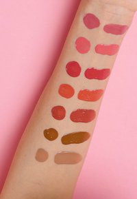 Muestras de prueba de varios colores de labios en un brazo con tono de piel, mostrando tonalidades desde rojo hasta marrón sobre un fondo rosa.