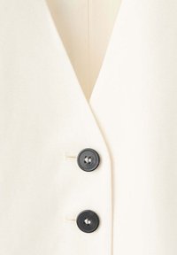 Gros plan d’un vêtement couleur crème avec deux boutons noirs et des boutonnières le long de l’ouverture avant.