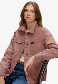 Różowa bawełniana kurtka z kołnierzykiem, zapięciem na guziki, wieloma kieszeniami z przodu oraz naszywką na piersi, w połączeniu z dżinsami i plecionym paskiem.