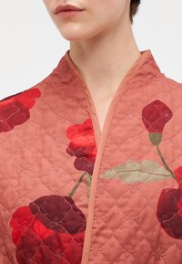 Quiltet jakke i ferskenfarvet stof med store røde blomsterdesign. Designet har en sjalkrave og dekorative syninger.